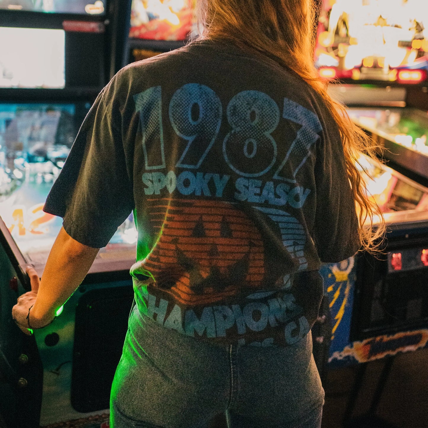 Person wearing a t-shirt with '1988 Spooky Season Champion' text in an arcade setting