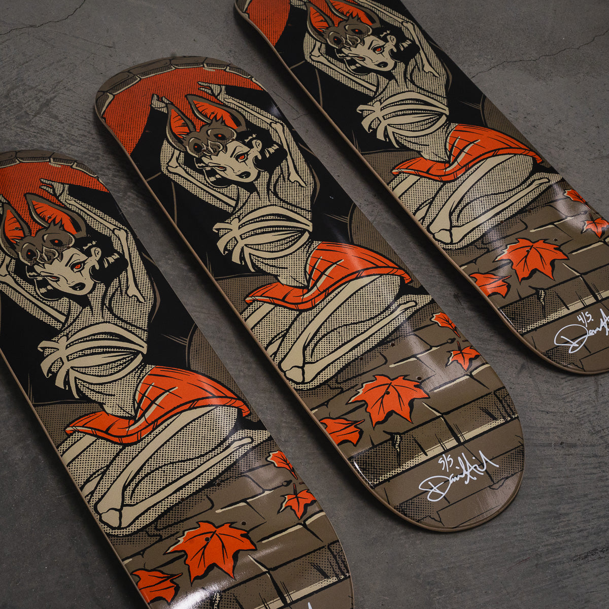 Three skateboard decks arranged diagonally on a dark gray textured surface, featuring a vintage-style illustration of a woman in a kimono with orange leaves and brick-patterned details.