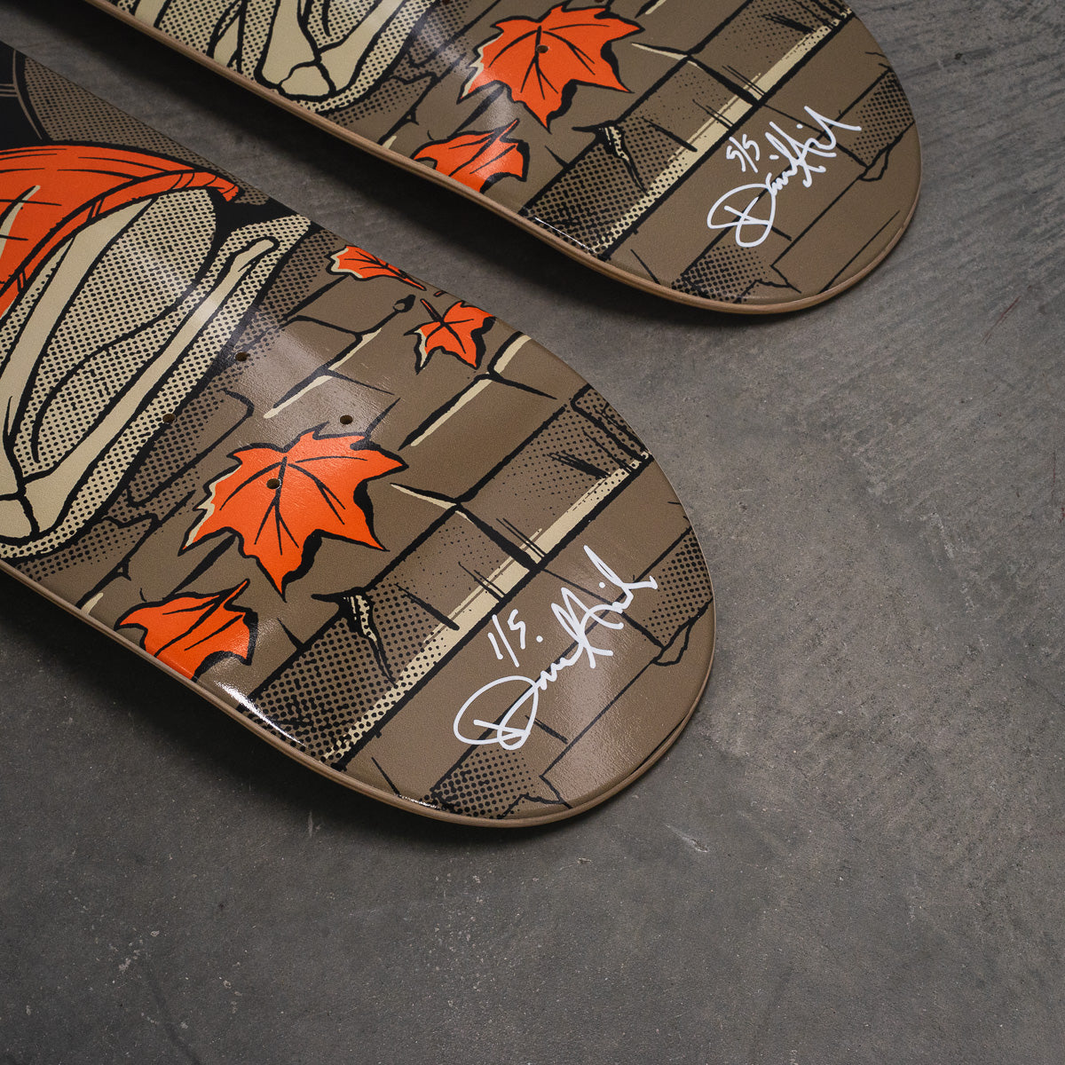 Two skateboard decks laid diagonally on a dark gray concrete floor, featuring a graphic design with orange maple leaves, a brick wall, and white script signatures.