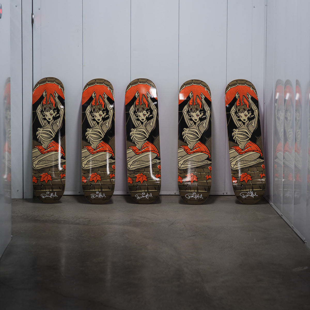Five skateboard decks standing upright in a row against a white paneled wall, featuring a graphic design of a kneeling woman with red hair and a brick-pattern background.