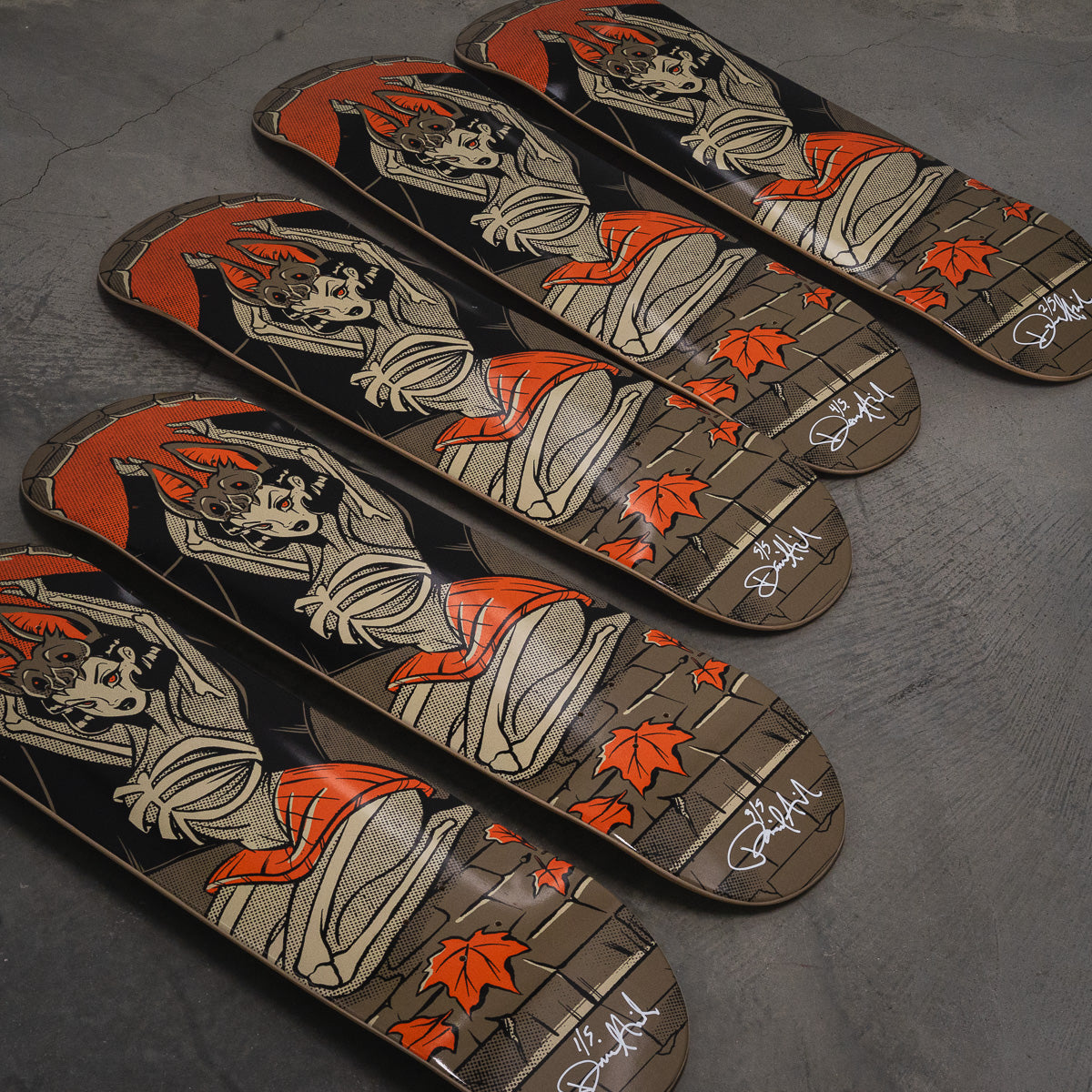 Four skateboard decks arranged diagonally on a dark gray concrete floor, featuring a vintage-style illustration of a woman in a kimono with red hair, orange maple leaves, and a brick-pattern background. The decks have white signatures at the nose.