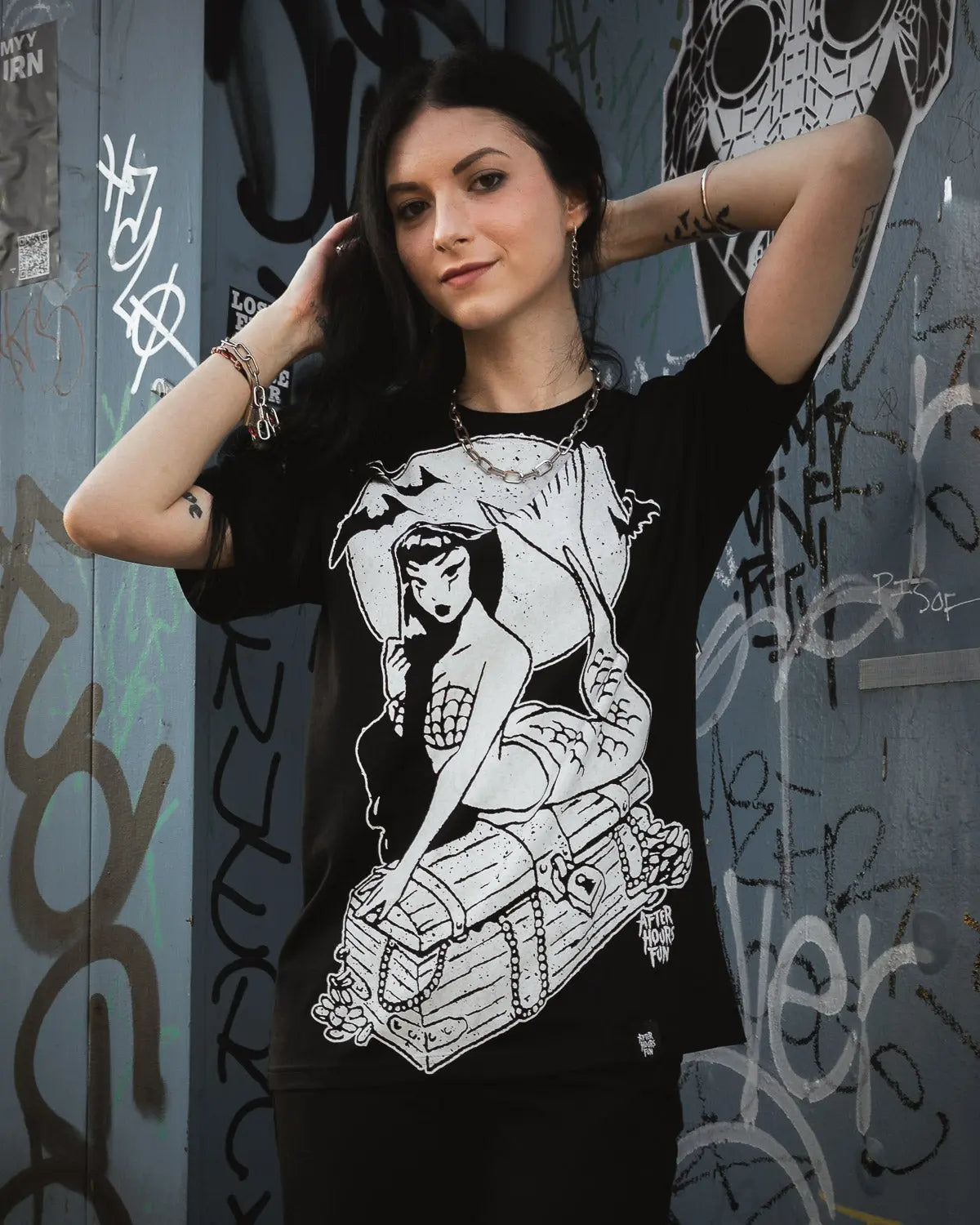 Woman wearing a black t-shirt with a graphic design of a woman on a box, standing against a graffiti-covered wall.